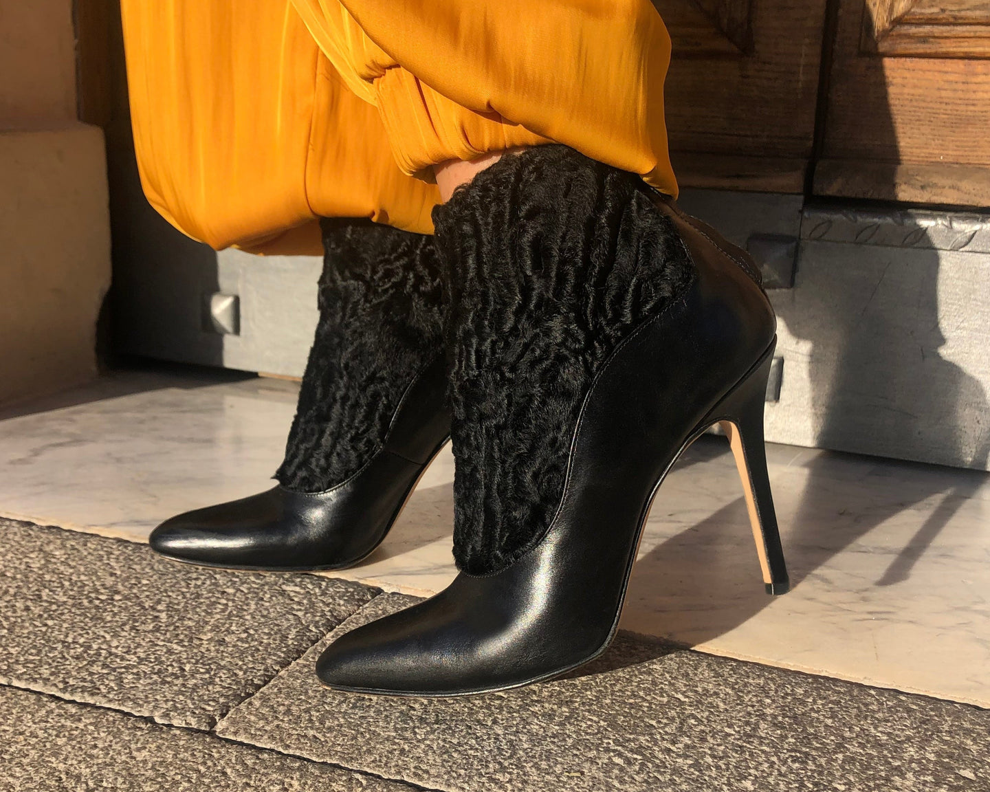Black high-heeled shoes with textured design worn by a person in mustard pants on a stone pavement.