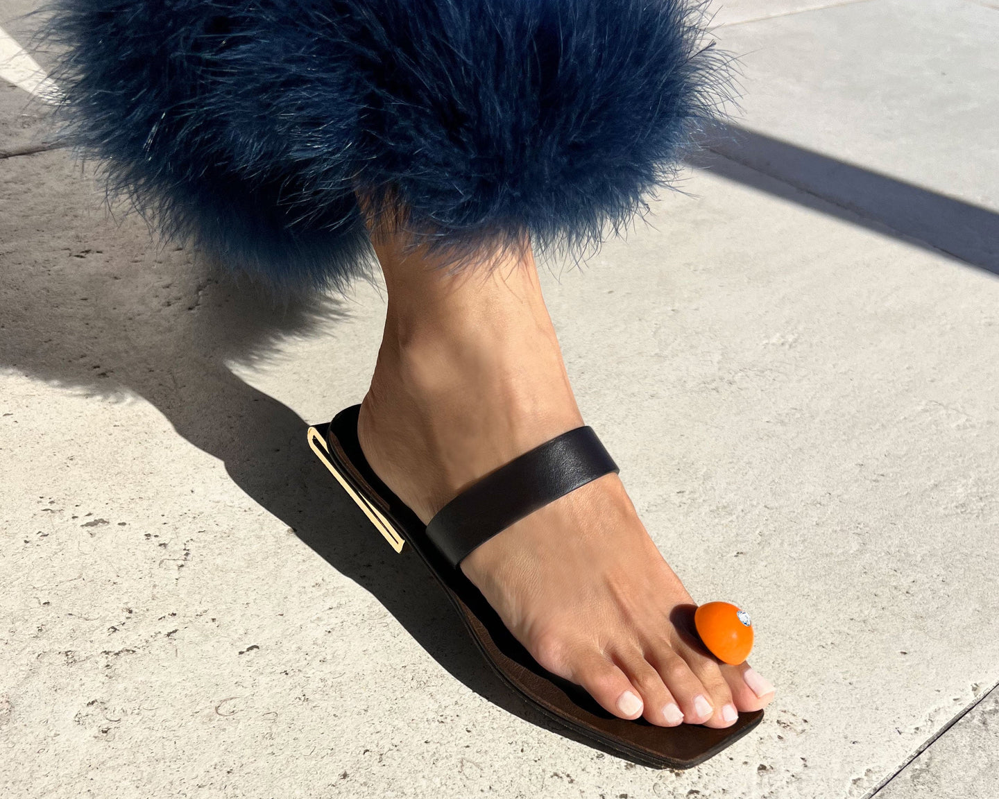 Black sandal with blue feathers on a foot, standing on a concrete surface.